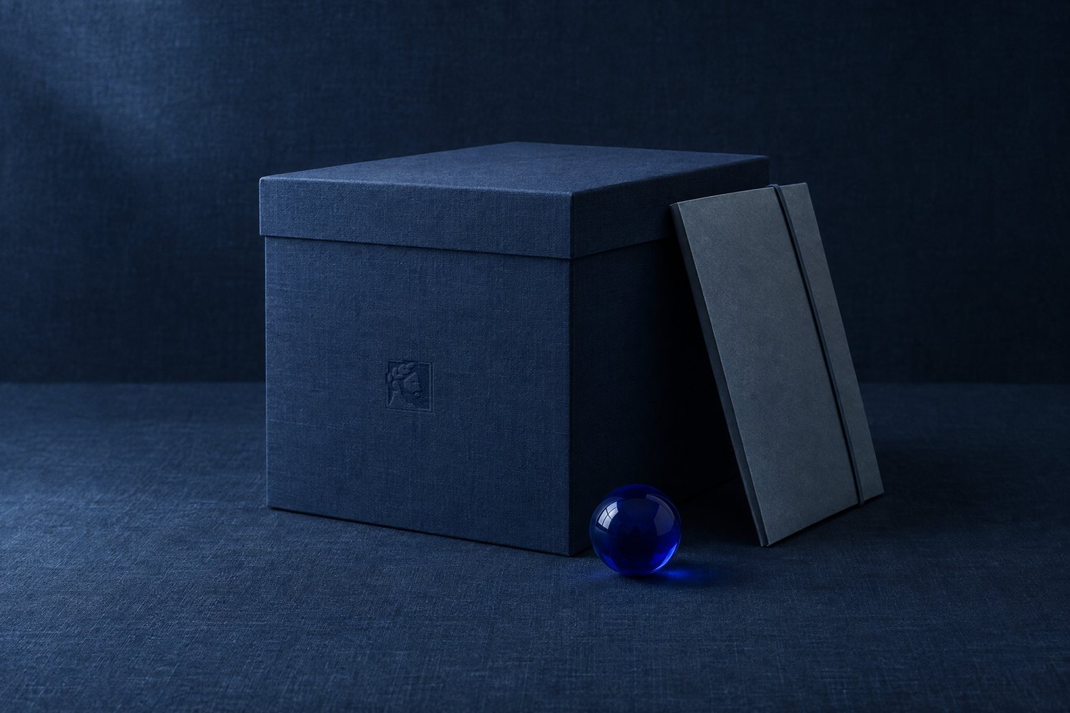 A navy linen archive box with a slate folder leaning on it and a cobalt glass sphere, the Olivent mark pressed into the box front