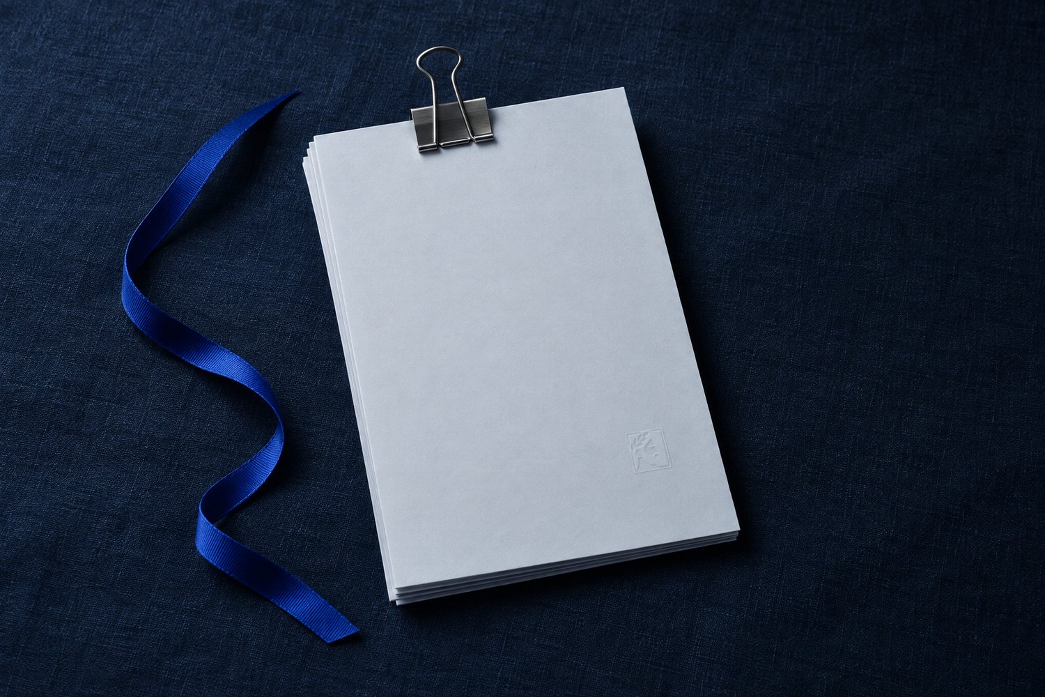 A stack of blank pages held by a steel binder clip, the Olivent mark embossed in the corner, a cobalt ribbon beside it