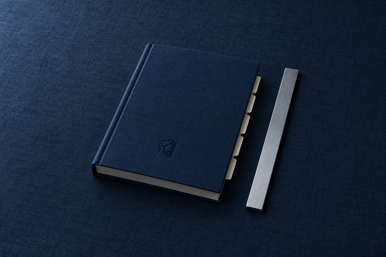 A closed navy clothbound ledger with cream tab dividers and a steel straightedge, the Olivent mark pressed into the cover