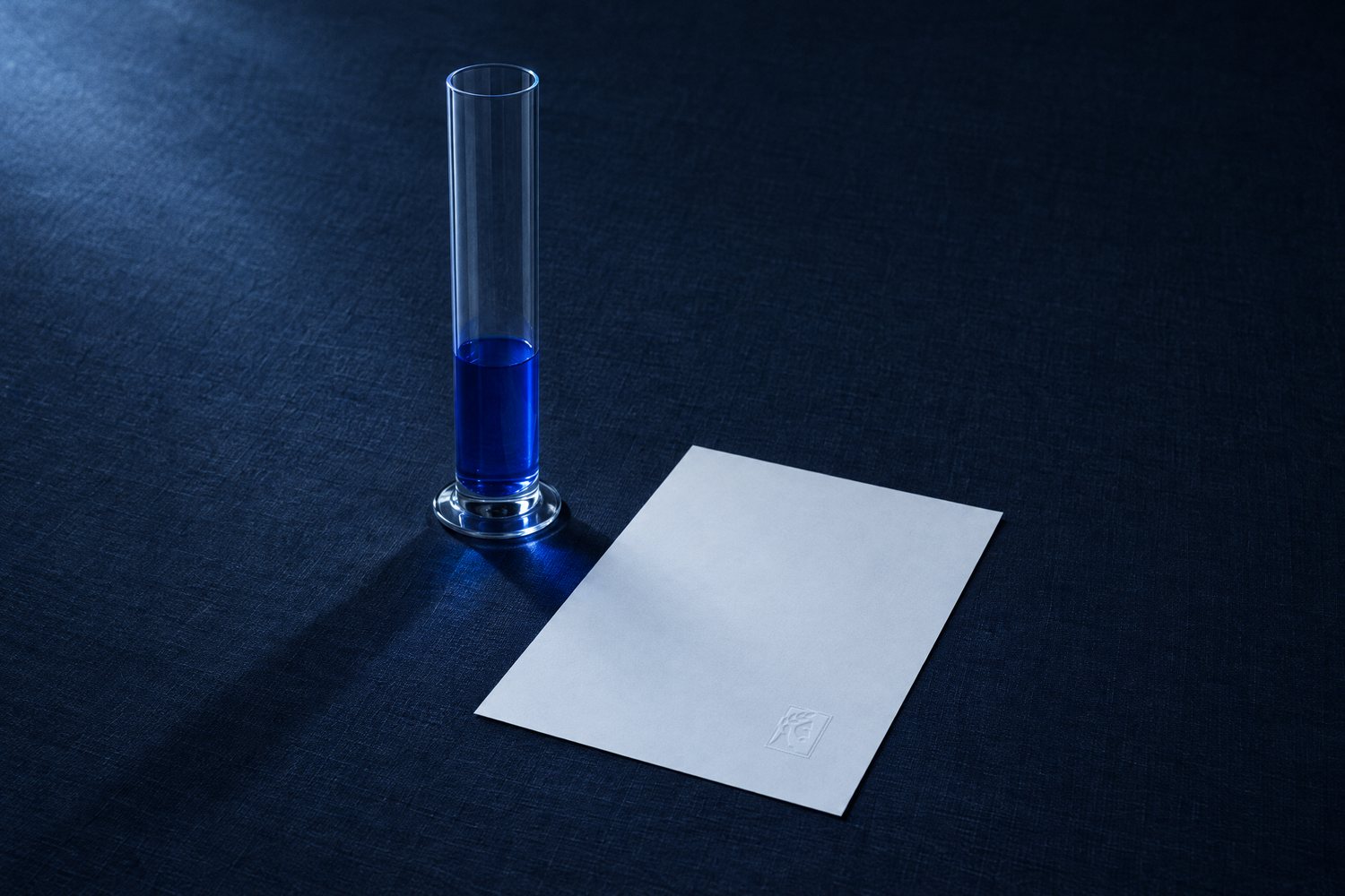 A tall glass cylinder a third full of cobalt liquid beside a blank sheet with the Olivent mark embossed at the corner