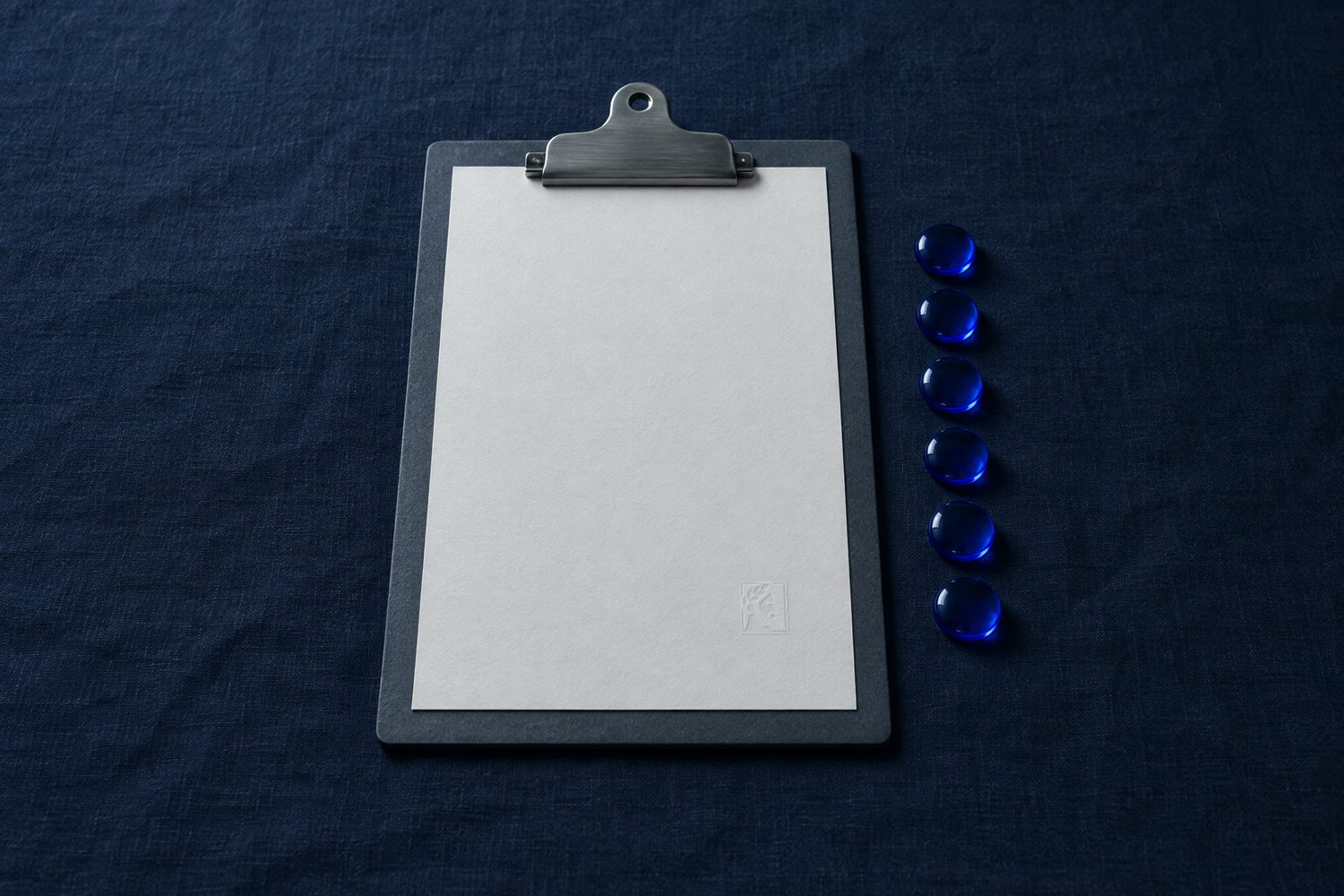 A blank sheet on a slate clipboard beside a column of six cobalt glass tokens, the Olivent mark embossed at the sheet's lower right