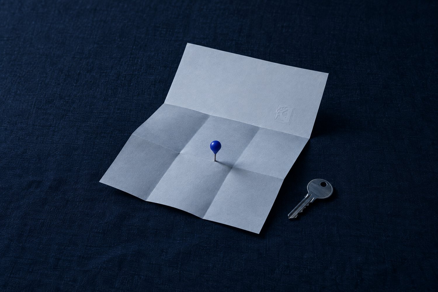 A blank folded paper map with a single cobalt pin and a steel house key, the Olivent mark embossed on the top panel