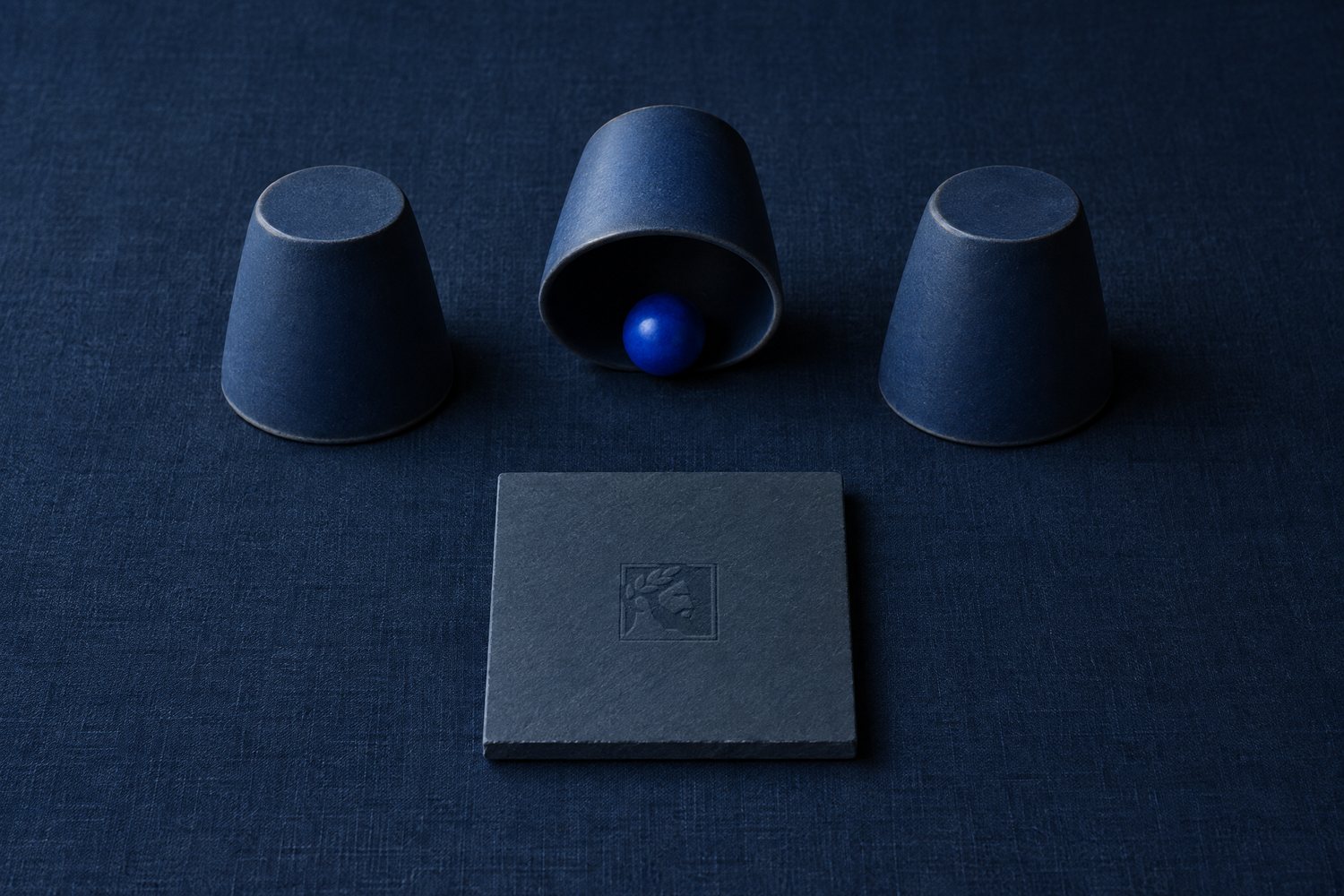 Three upturned navy cups, the middle one lifted to show a cobalt ball, and a slate tile debossed with the Olivent mark