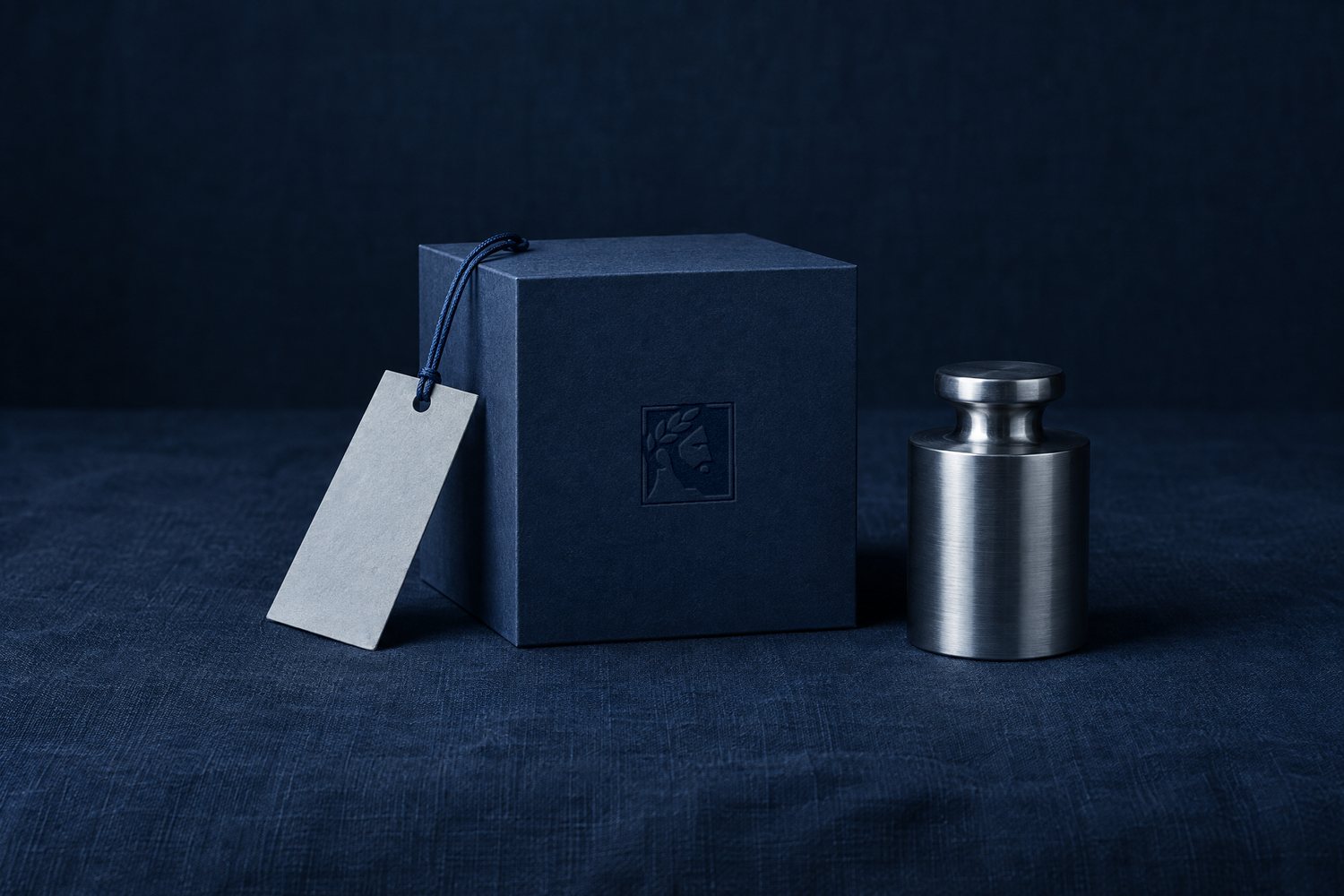 A navy cube debossed with the Olivent mark, a blank paper tag on a cord, and a brushed steel weight