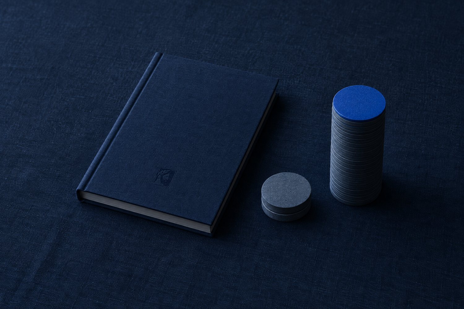 A closed navy cloth ledger with the Olivent mark pressed into its cover, beside a short stack of two slate discs and a tall stack topped with a cobalt disc